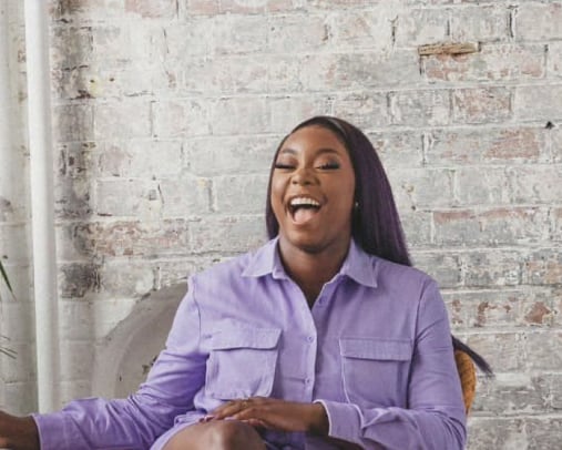 Photo of Creative Access mentee, Jay McGregor. Jay is a Black woman wearing a purple jumpsuit. She has her eyes shut and is laughing whilst sat down.